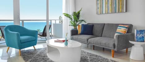 Un moderno salón con un sofá gris, un sillón azul, una mesa de centro blanca y vistas al océano.