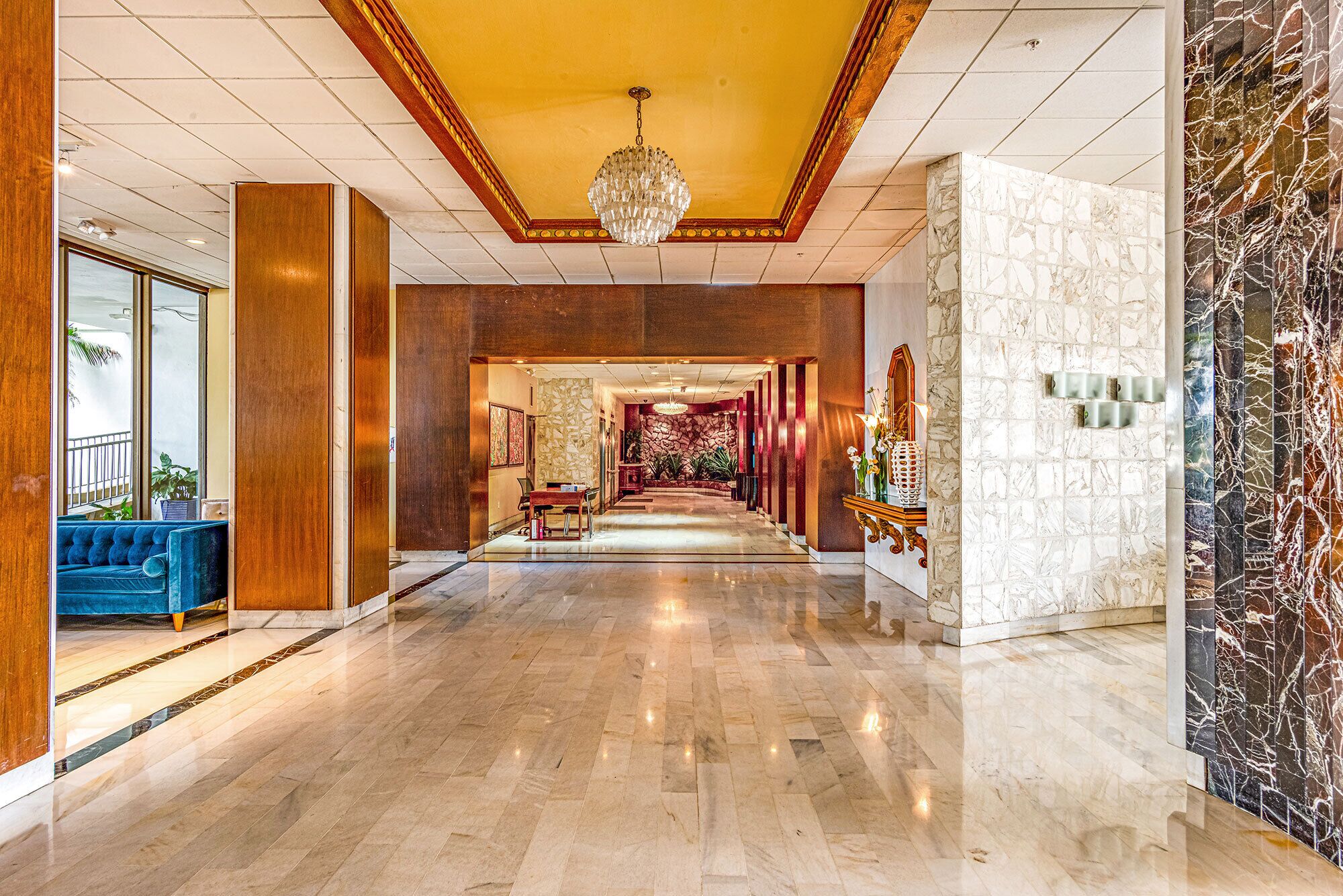 A spacious hallway with a marble floor, a chandelier, and a blue sofa.
