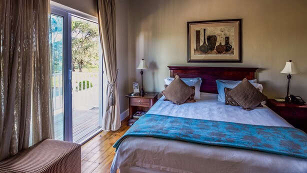 A hotel room with a large bed, a bench, a nightstand, a lamp, and a painting on the wall. - A&A Guesthouse (Gqeberha (Port Elizabeth))