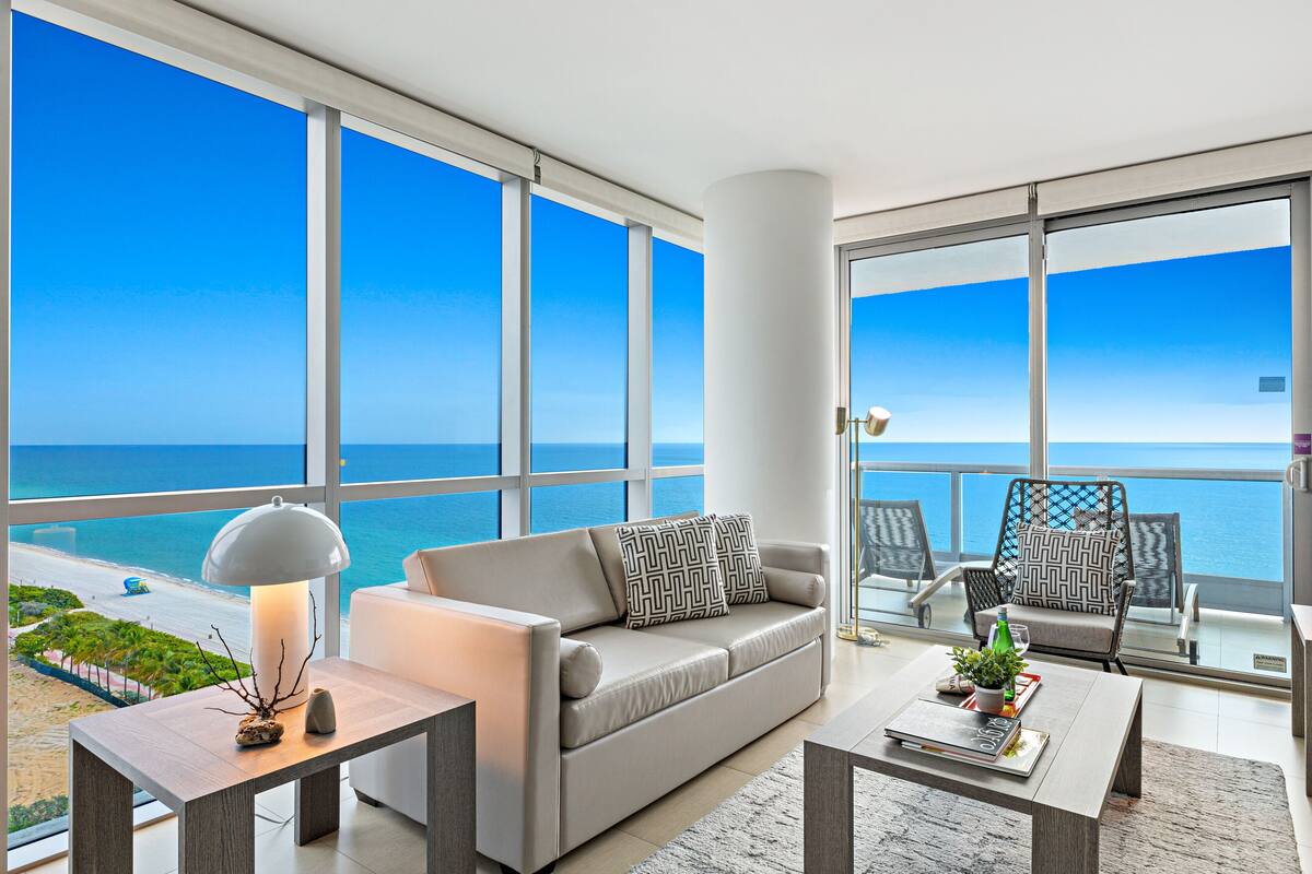 A modern living room with a large window overlooking the ocean, a sofa, a coffee table, and a balcony with chairs.