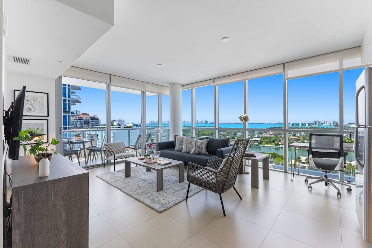 A spacious living room with a large window offering a city view, a sofa, a coffee table, and a dining area.