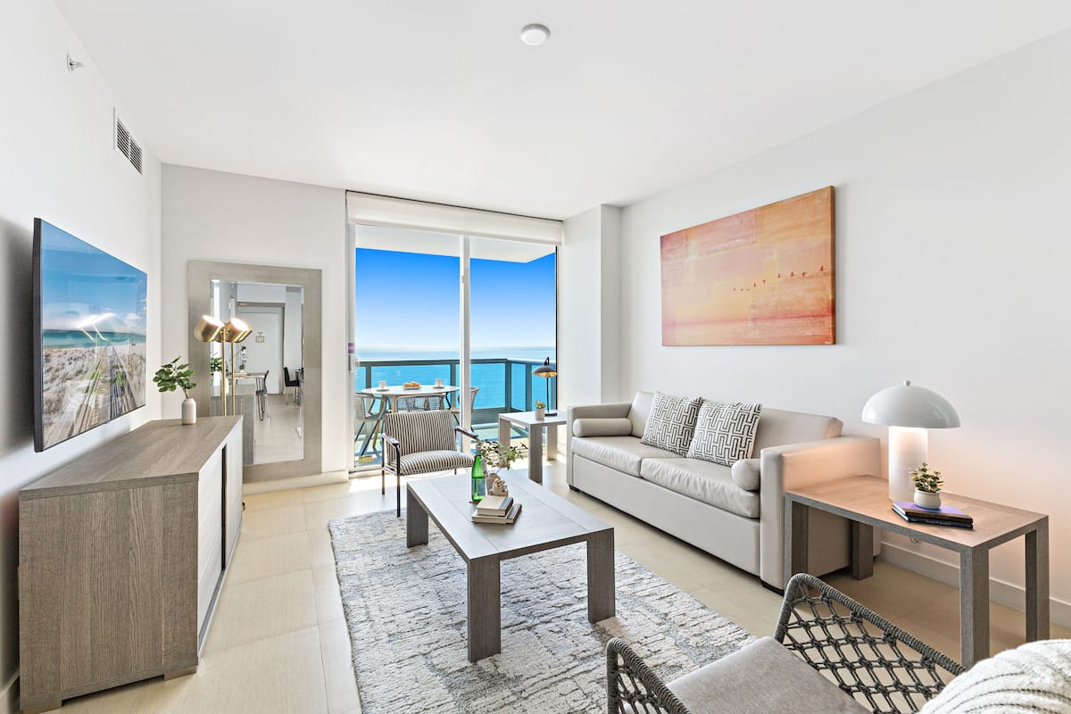 A modern living room with a sofa, coffee table, and chairs, a TV, and a large window with a view of the sea.