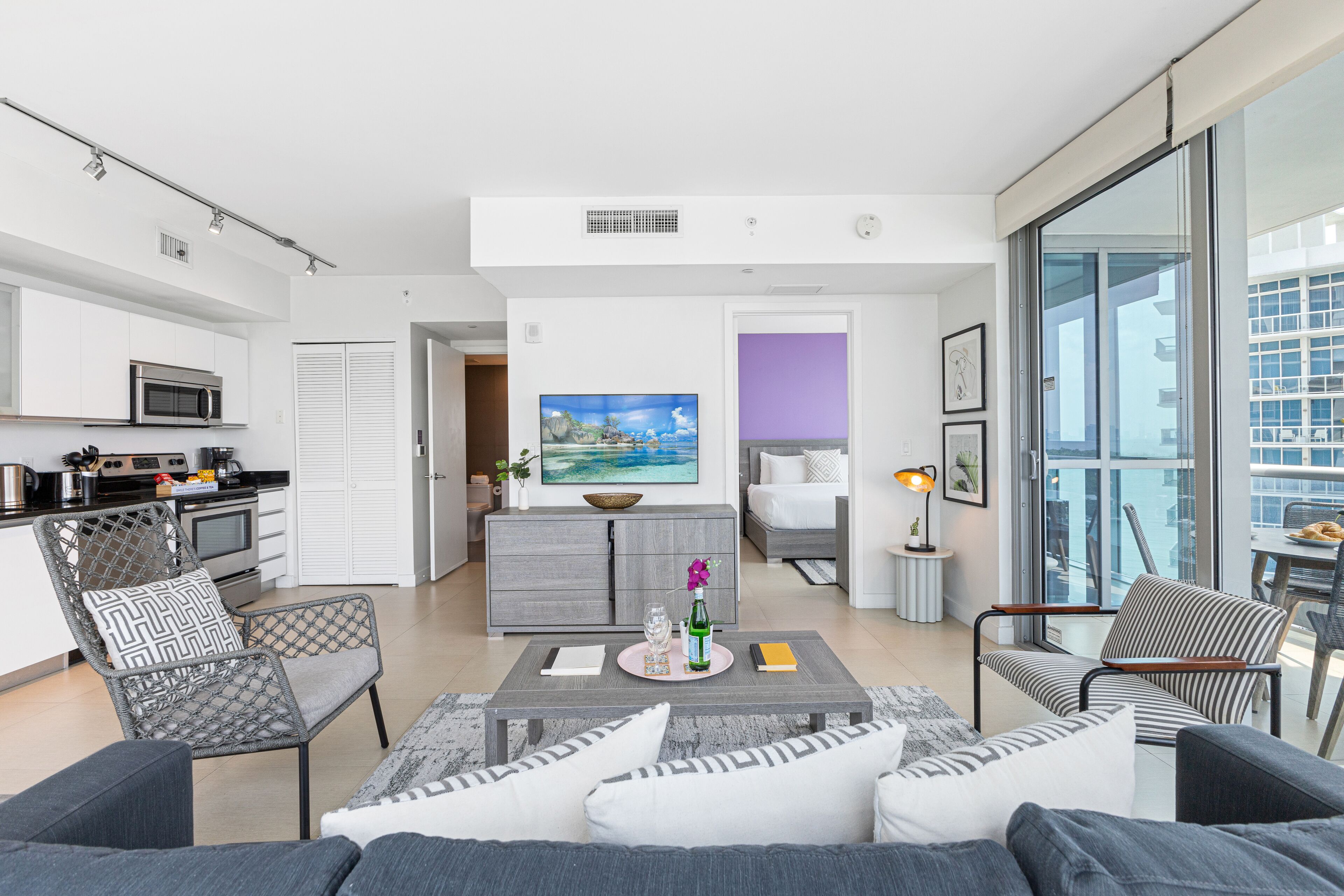 Un apartamento moderno con cocina, comedor y sala de estar. Cuenta con un ventanal grande, un televisor de pantalla plana y un balcón con vistas a la ciudad.