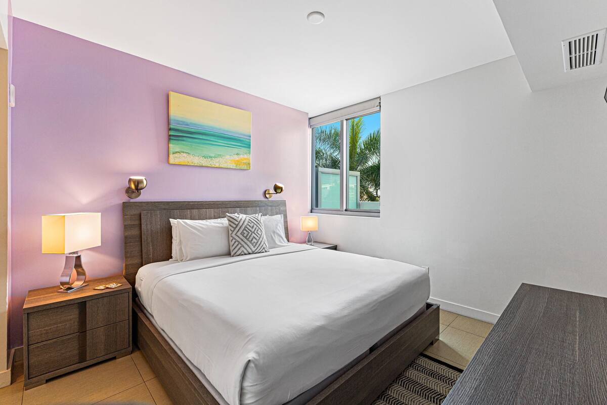 A modern hotel room with a large bed, two bedside tables with lamps, a painting of a beach scene, and a window with a view of palm trees.