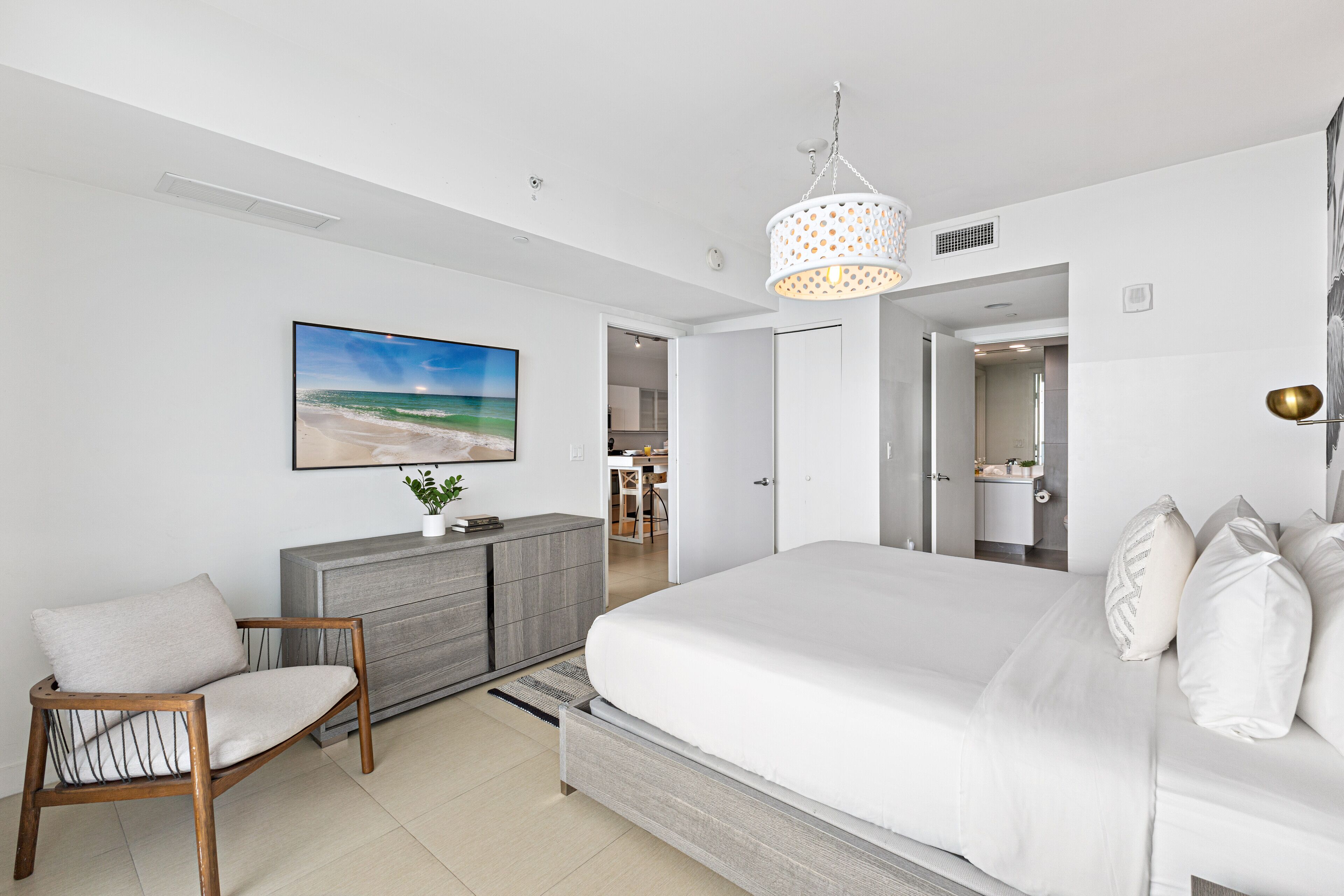 A modern bedroom with a large bed, a wooden chair, a grey dresser, and a painting of a beach scene.