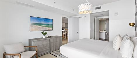 A modern bedroom with a large bed, a wooden chair, a grey dresser, and a painting of a beach scene.