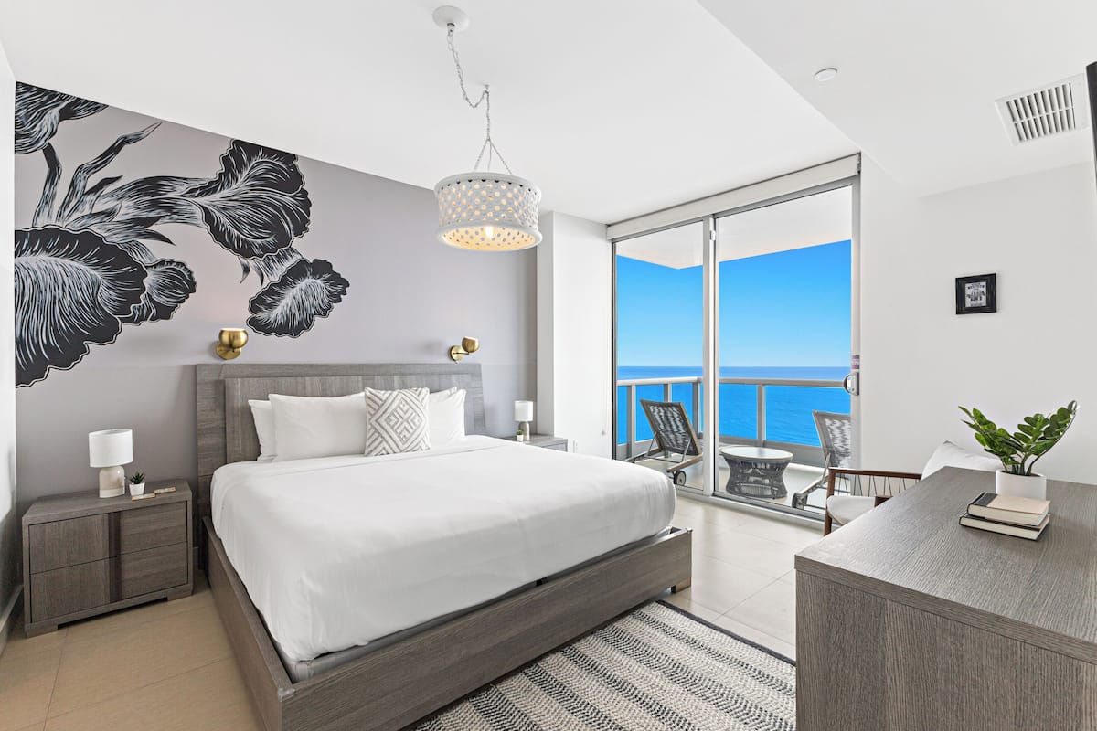 A bedroom with a large bed, a nightstand, a desk with a plant, and a view of the sea through a glass door.