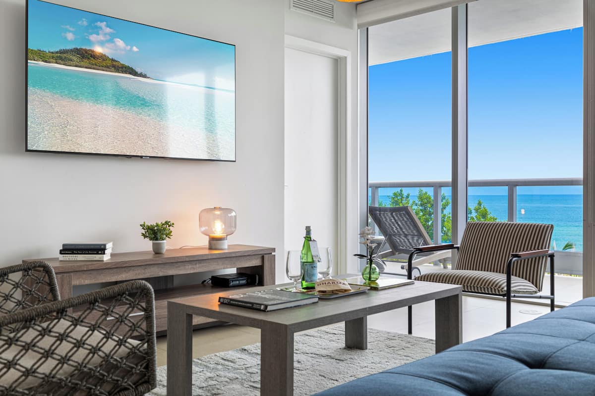A modern living room with a large TV, a wooden coffee table, and two chairs. There is a view of the ocean through large windows.