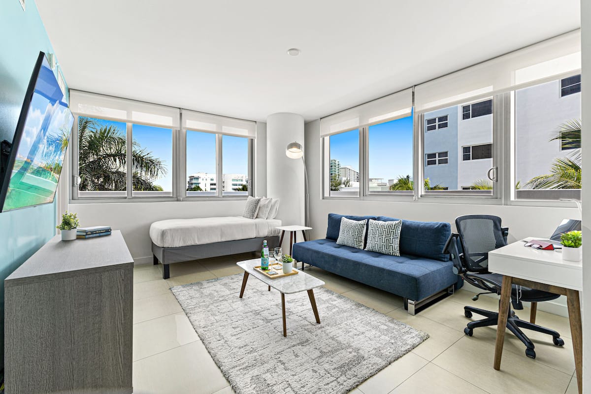 A modern living room with a sofa, a desk, and a coffee table. There are large windows providing a view of the city.