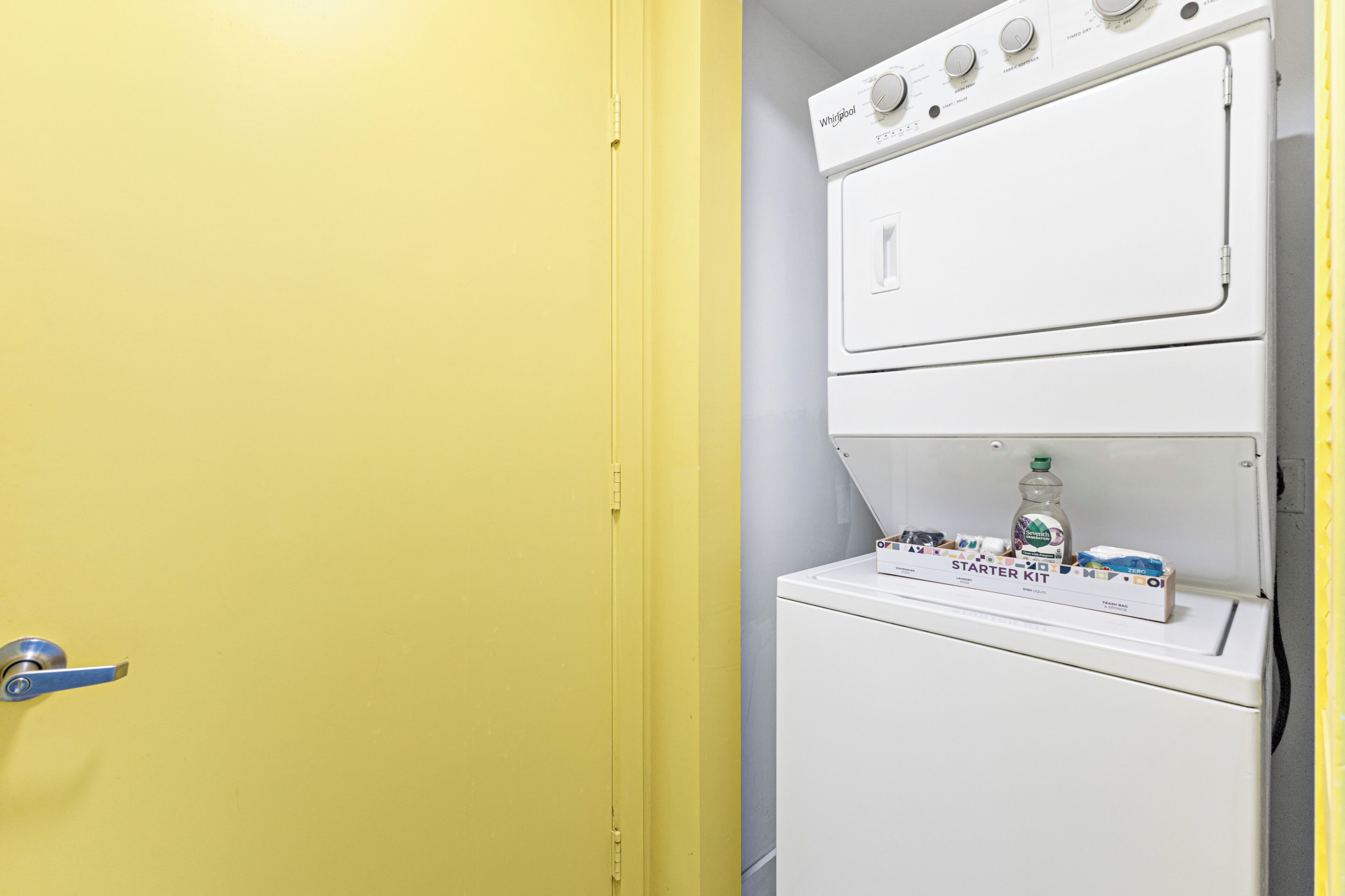 Een wasserij met een gele deur, een wasmachine en droger boven elkaar, en een 'STARTER KIT' op de droger.