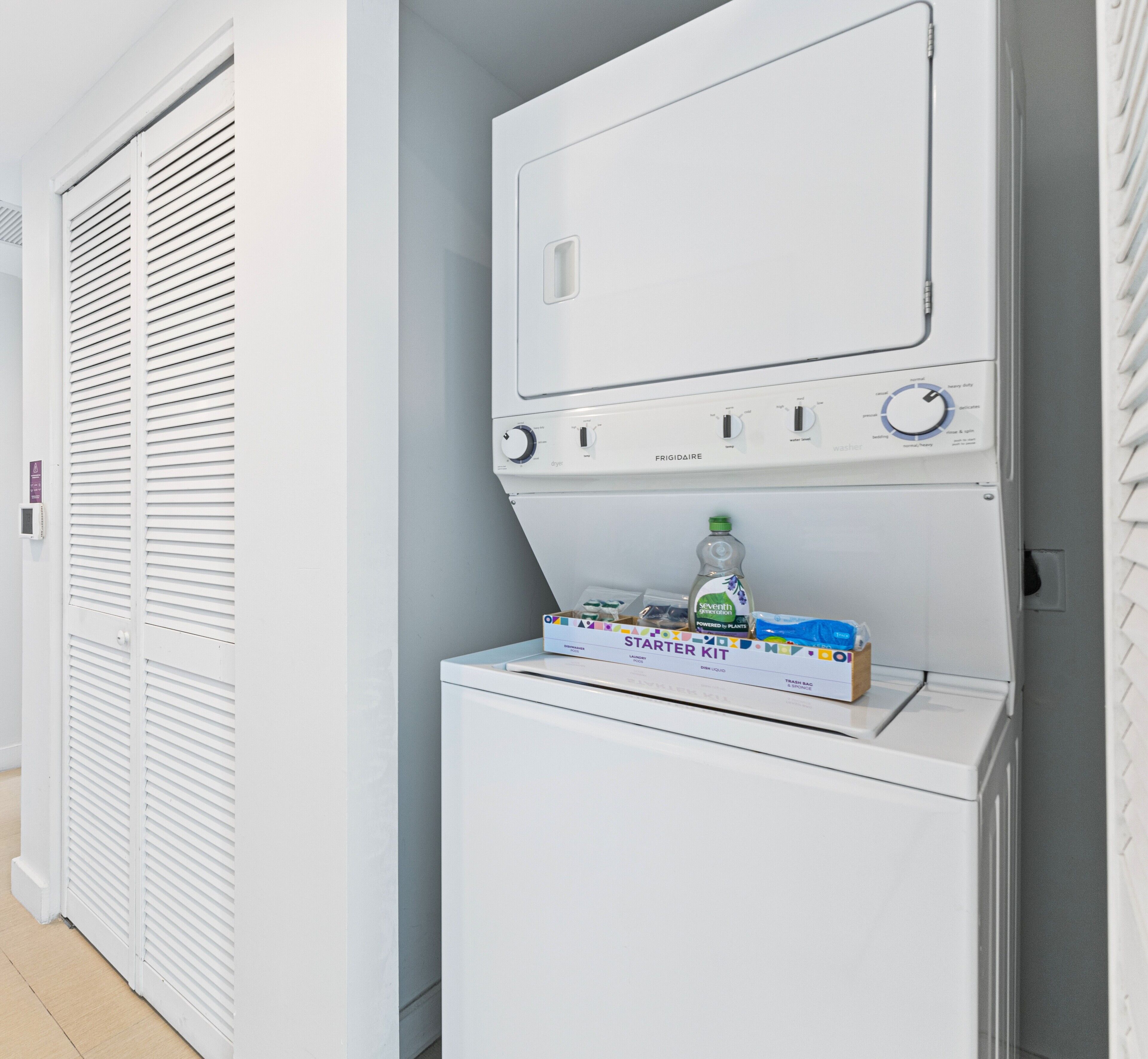A compact, white laundry machine with a control panel and a small shelf holding a bottle and a laundry kit.