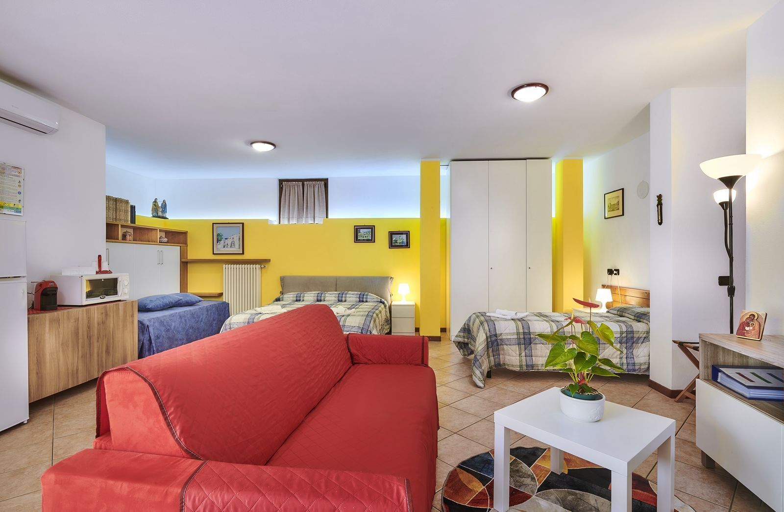 A compact living space with a kitchenette, a red sofa, two beds with different bedding, a small table with a plant, and a ceiling light.