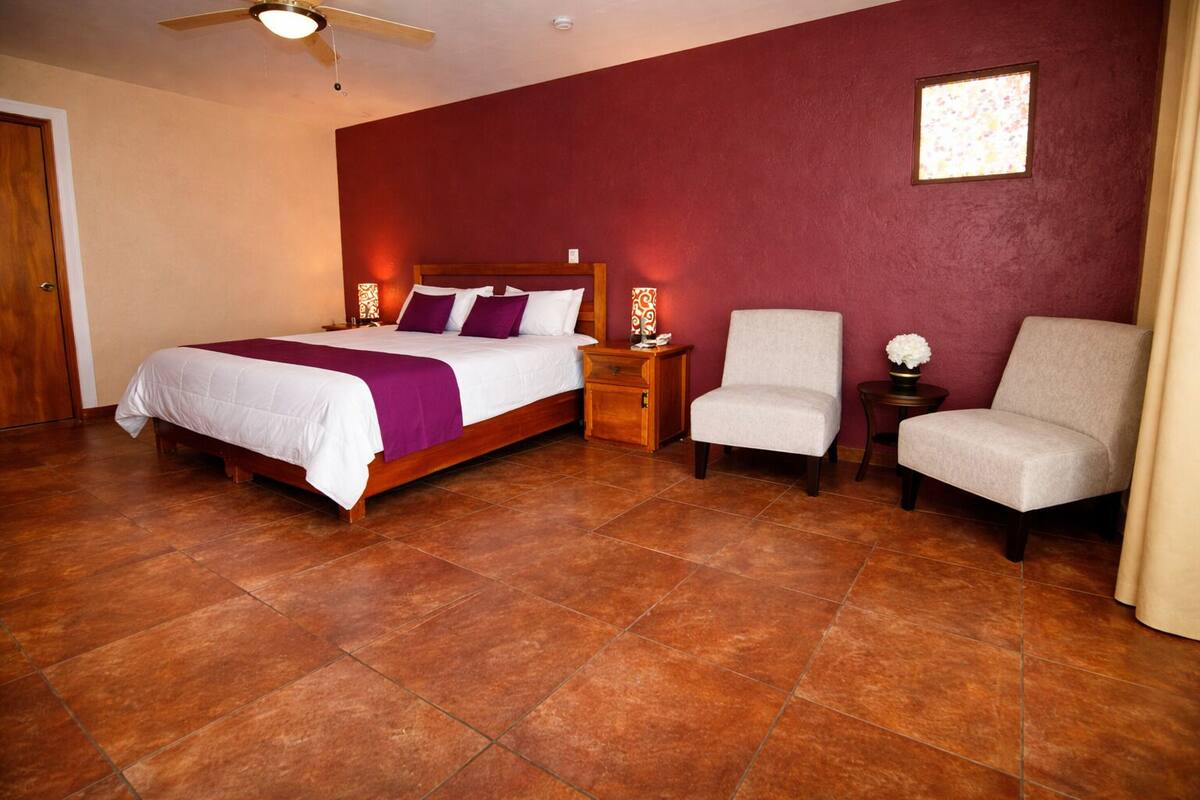 A hotel room with a bed, two armchairs, a nightstand, a wooden headboard, a ceiling fan, and a window with a view.