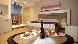 Ein Hotelzimmer mit Bett, Nachttisch, einer Weinflasche, Obstschale und einem Frühstücksteller mit Croissants.
