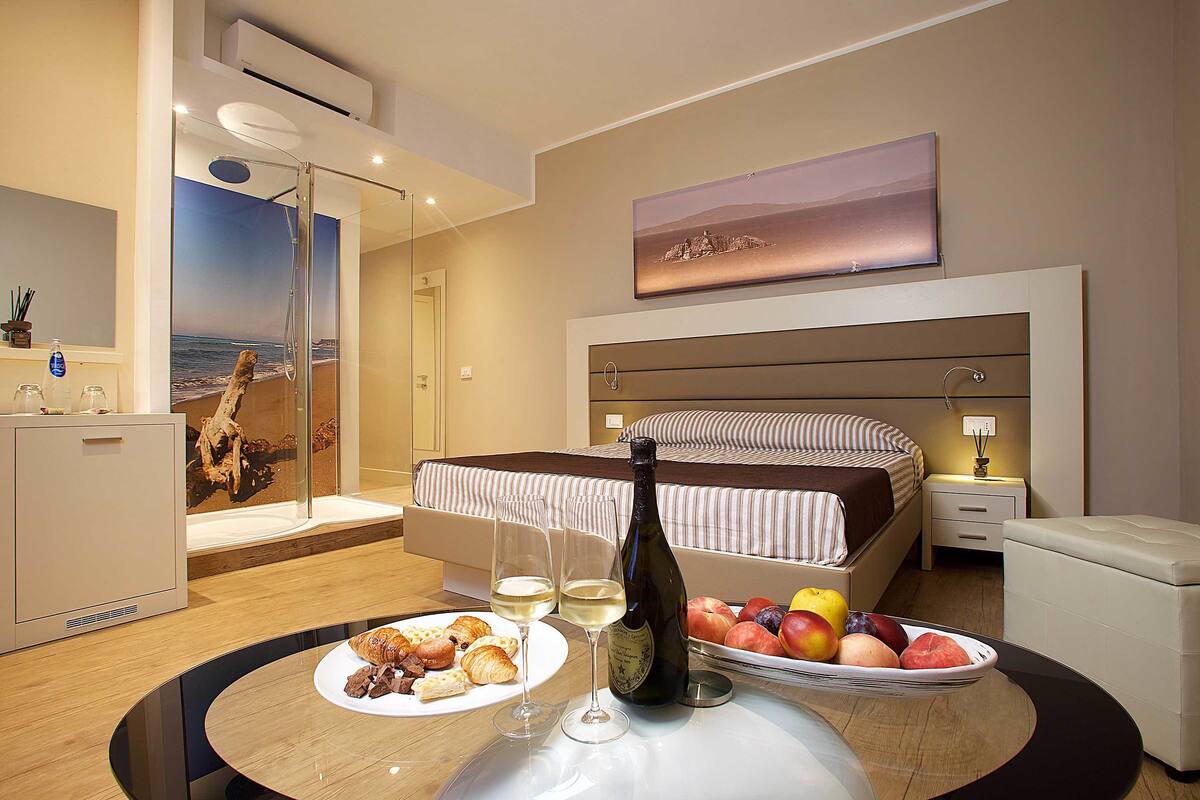 Ein Hotelzimmer mit Bett, Nachttisch, einer Weinflasche, Obstschale und einem Frühstücksteller mit Croissants.