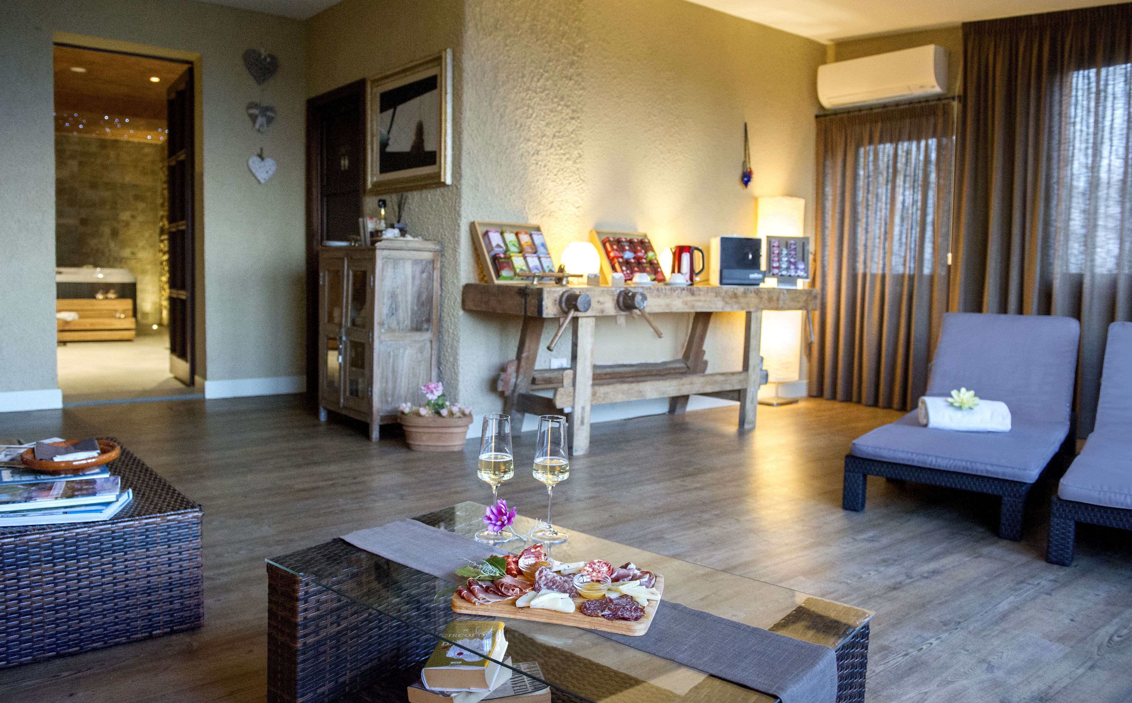 Een woonkamer met een houten salontafel waarop een vleesplank, twee wijnglazen en een potplant staan. Er is een rieten bank en een comfortabele fauteuil.