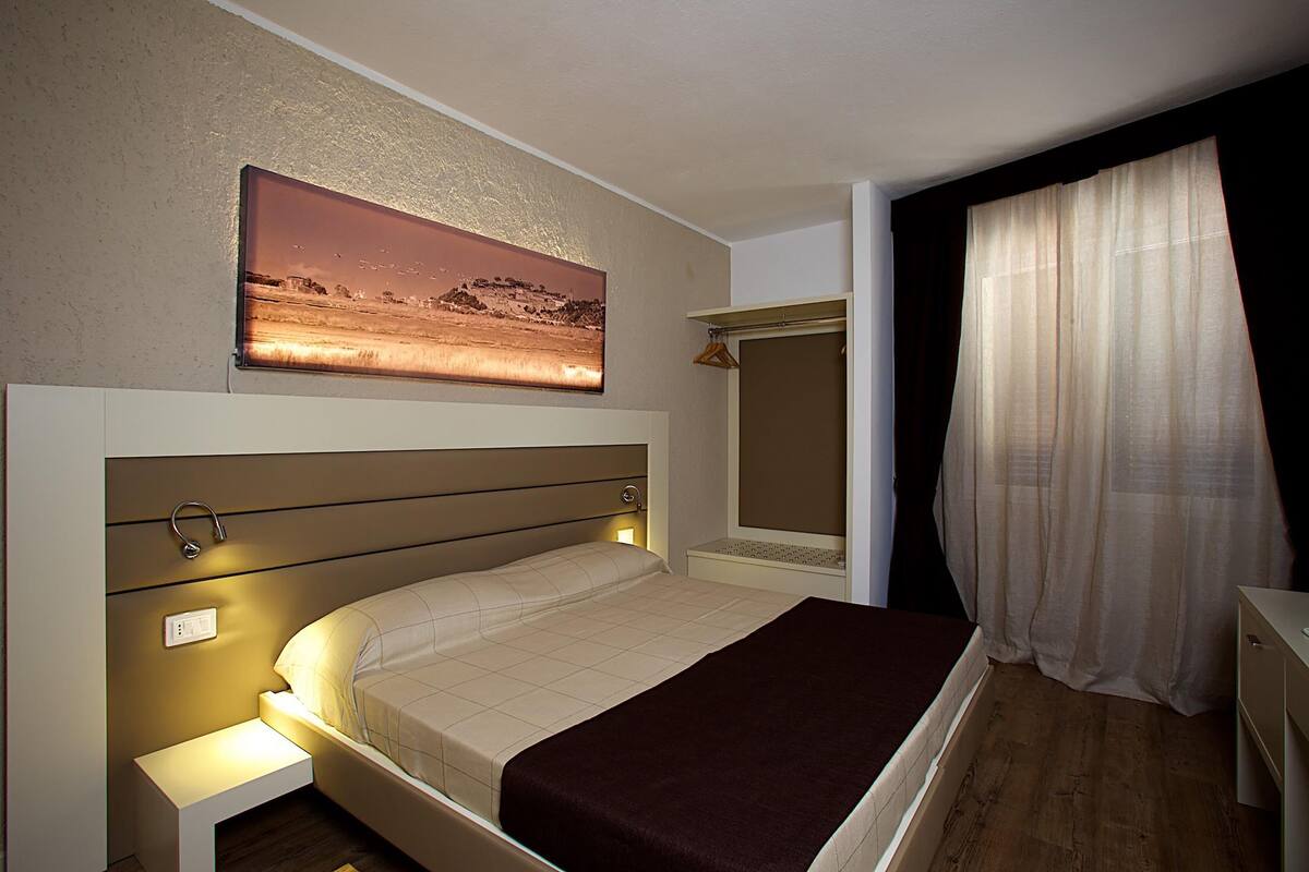 Ein Hotelzimmer mit Bett, Nachttisch, Wandbild und einem Fenster mit durchsichtigen Vorhängen.