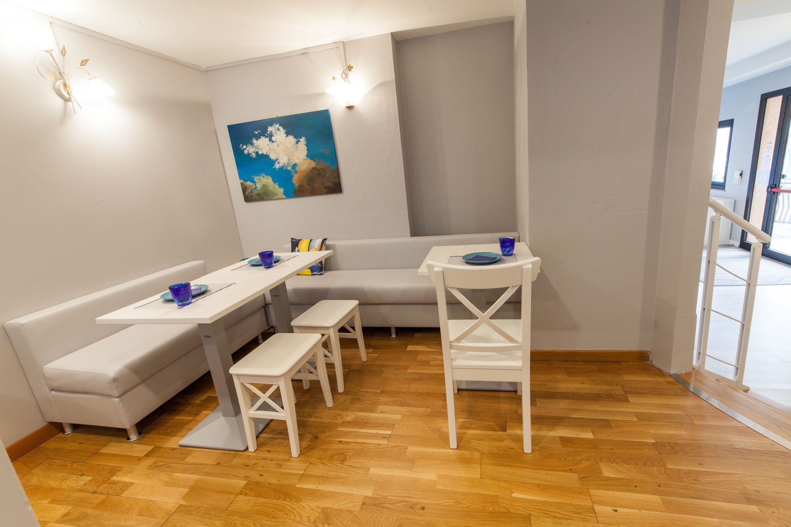 A modern, well-lit room with a wooden floor, a white sofa, a dining table with benches, and a painting on the wall.