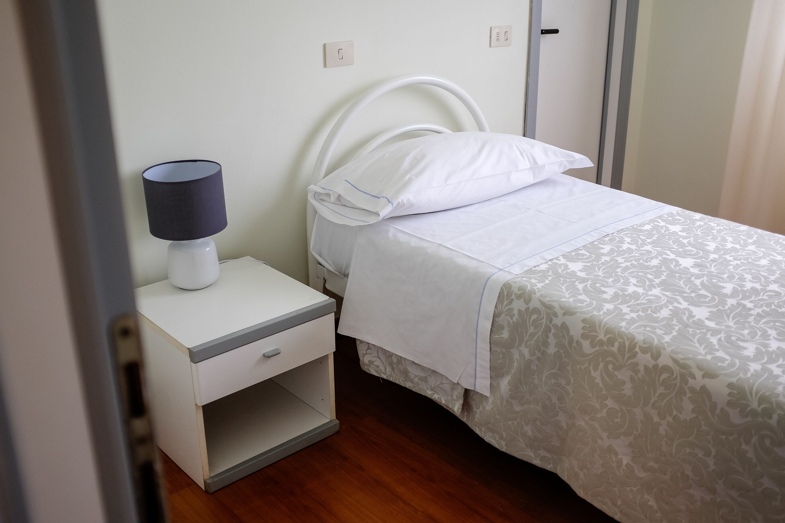 A neatly made bed with a patterned bedspread, a white bedside table with a lamp, and a small drawer.