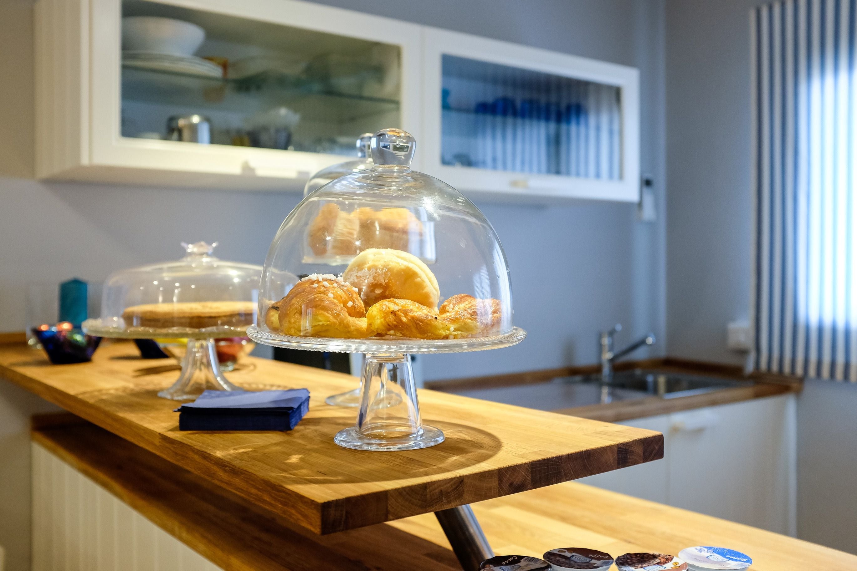 Una mesa de buffet con diversos pasteles bajo campanas de cristal, una encimera de madera y una zona de cocina al fondo.