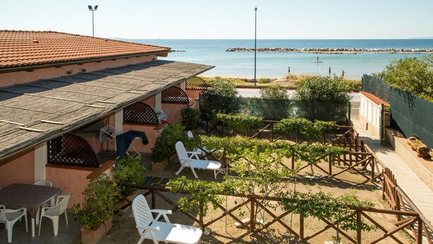 A patio with white chairs and a table, overlooking a beach with a rocky breakwater. - Le Dune (Follonica)