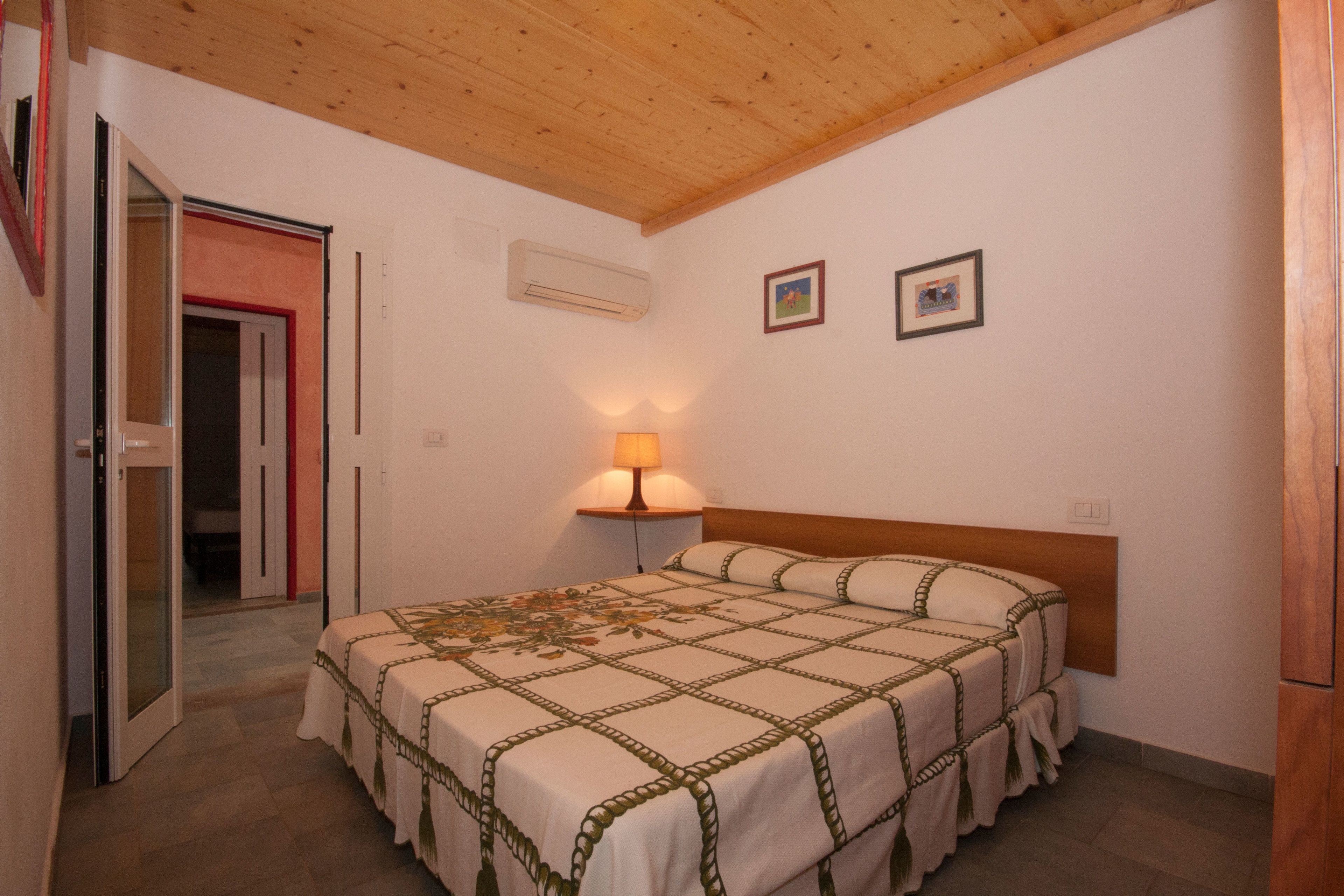 Une chambre avec un plafond en bois, un lit recouvert d’une couvre-lit à motifs, une lampe de chevet et deux tableaux encadrés accrochés au mur.