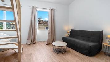 Une pièce avec un lit en mezzanine, un canapé noir, une table basse ronde et une fenêtre avec des rideaux.