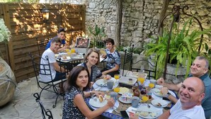 Eine Gruppe von Menschen speist im Freien auf einer Terrasse mit Steinmauern, Tischen, Stühlen und verschiedenen Speisen.