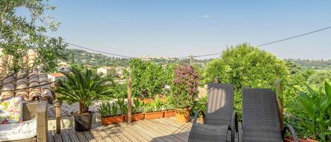 Une terrasse en bois, agrémentée de meubles en osier et de plantes en pot, qui offre une vue sur un paysage vallonné.