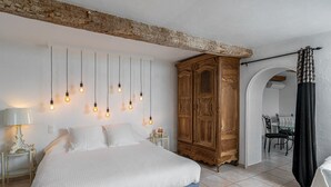 Ein Schlafzimmer mit einem großen Bett, hängenden Glühbirnen, einem Holzschrank und einem Essbereich, der durch einen Bogen sichtbar ist.