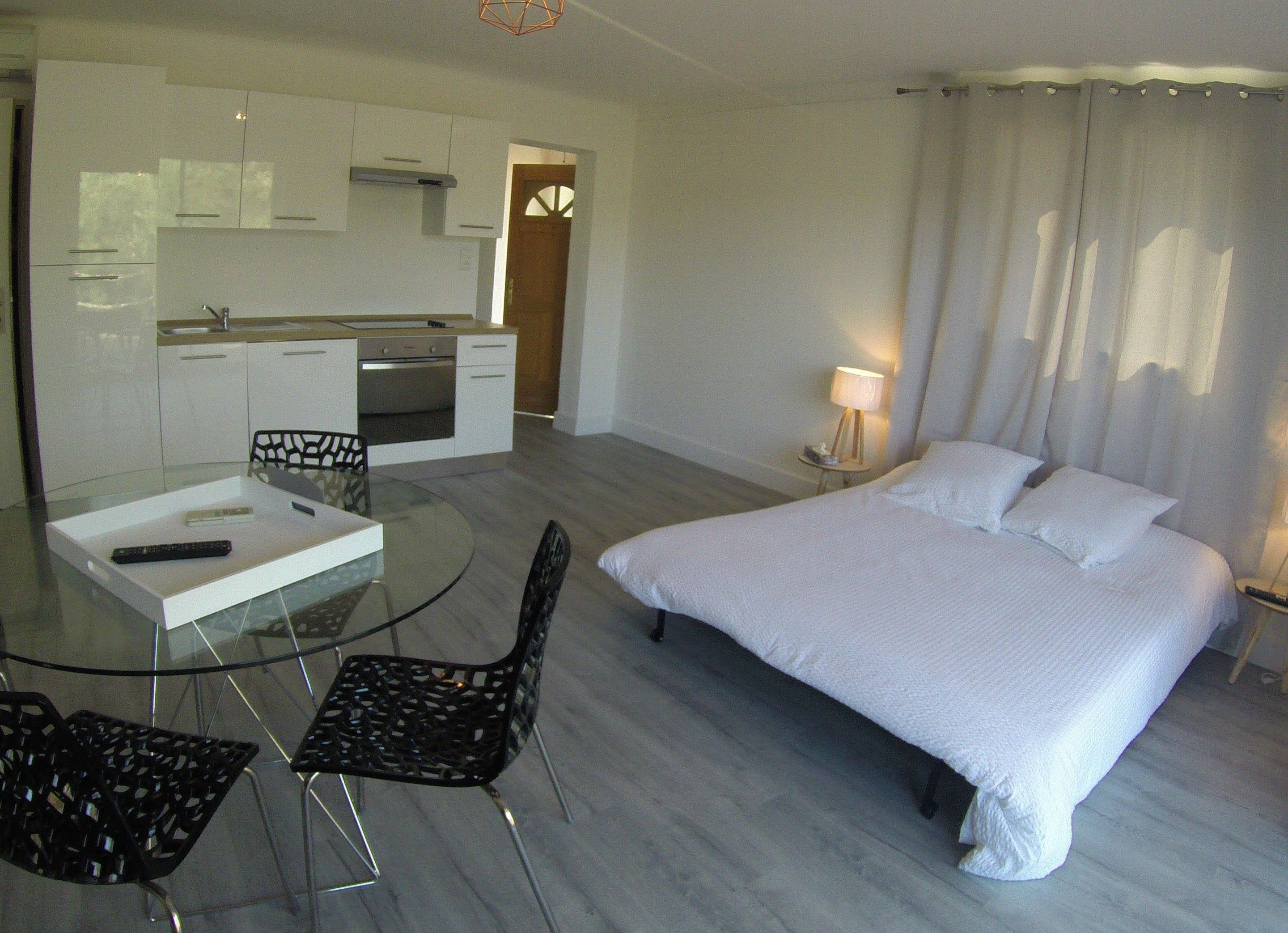 A modern hotel room with a glass dining table, two chairs, a bed with white linens, a small bedside table with a lamp, and a kitchen area in the background.