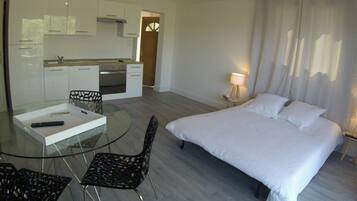 A modern hotel room with a glass dining table, two chairs, a bed with white linens, a small bedside table with a lamp, and a kitchen area in the background.