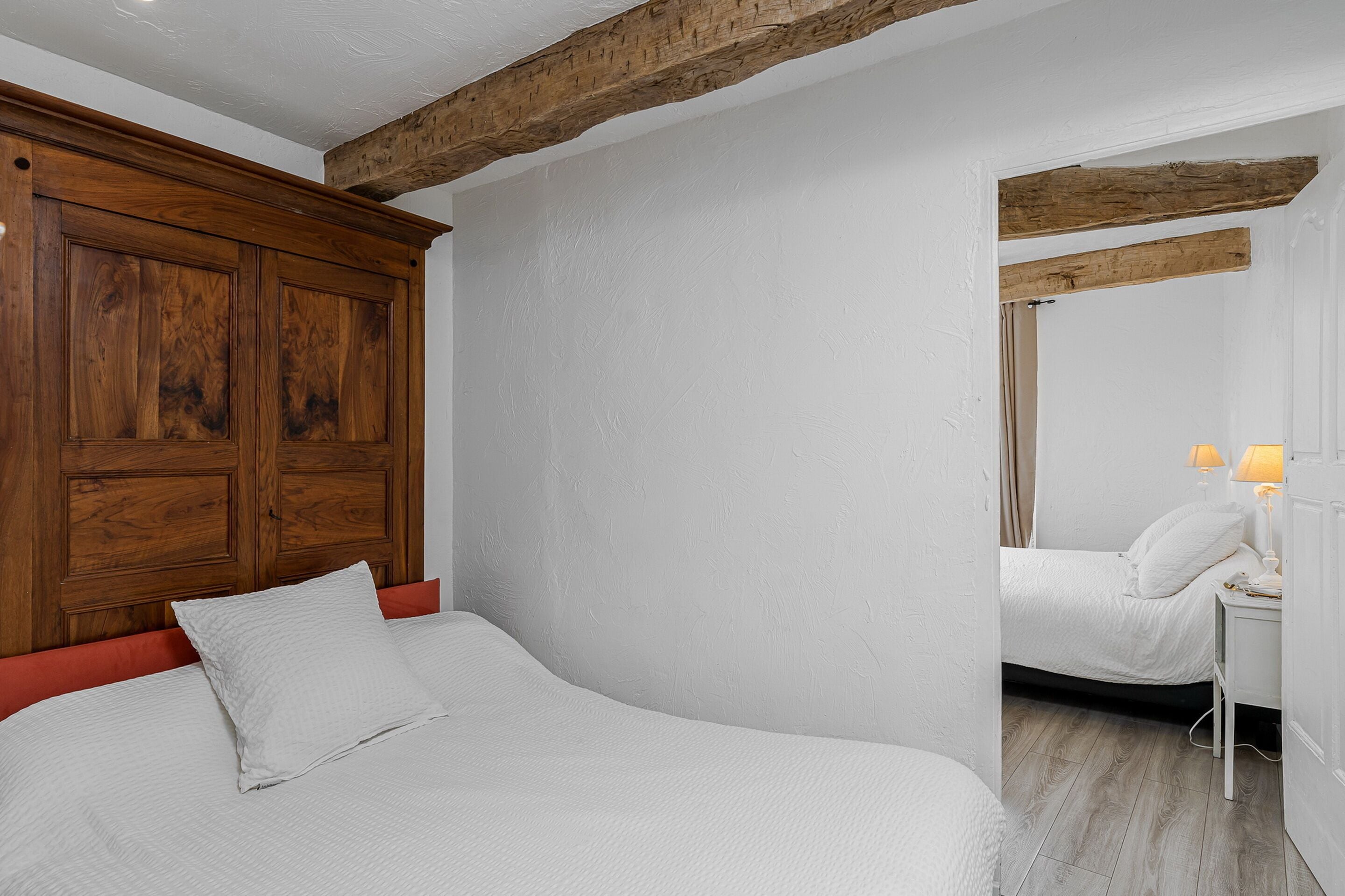 Un dormitorio con cabecera de madera, cama con ropa de cama blanca, mesita de noche con lámpara y suelo de madera.