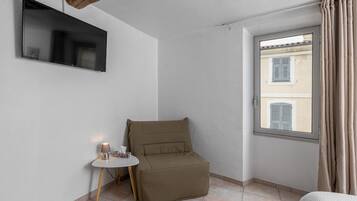 Una stanza piccola con una TV a schermo piatto montata a parete, una poltrona imbottita, un tavolino con una lampada e una finestra con tende.