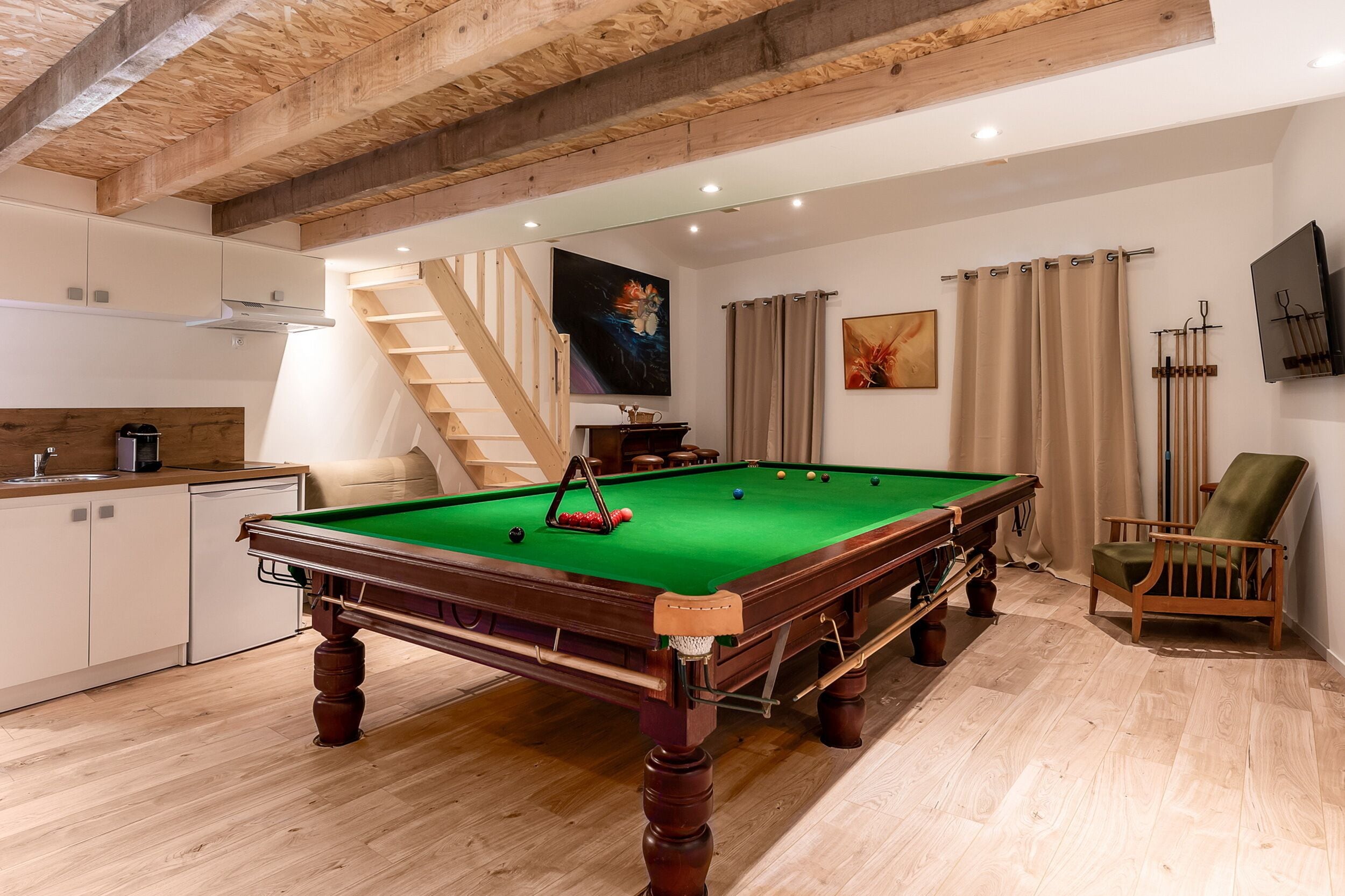 A pool table in a room with wooden floors, a staircase, a kitchen area, and a TV mounted on the wall.