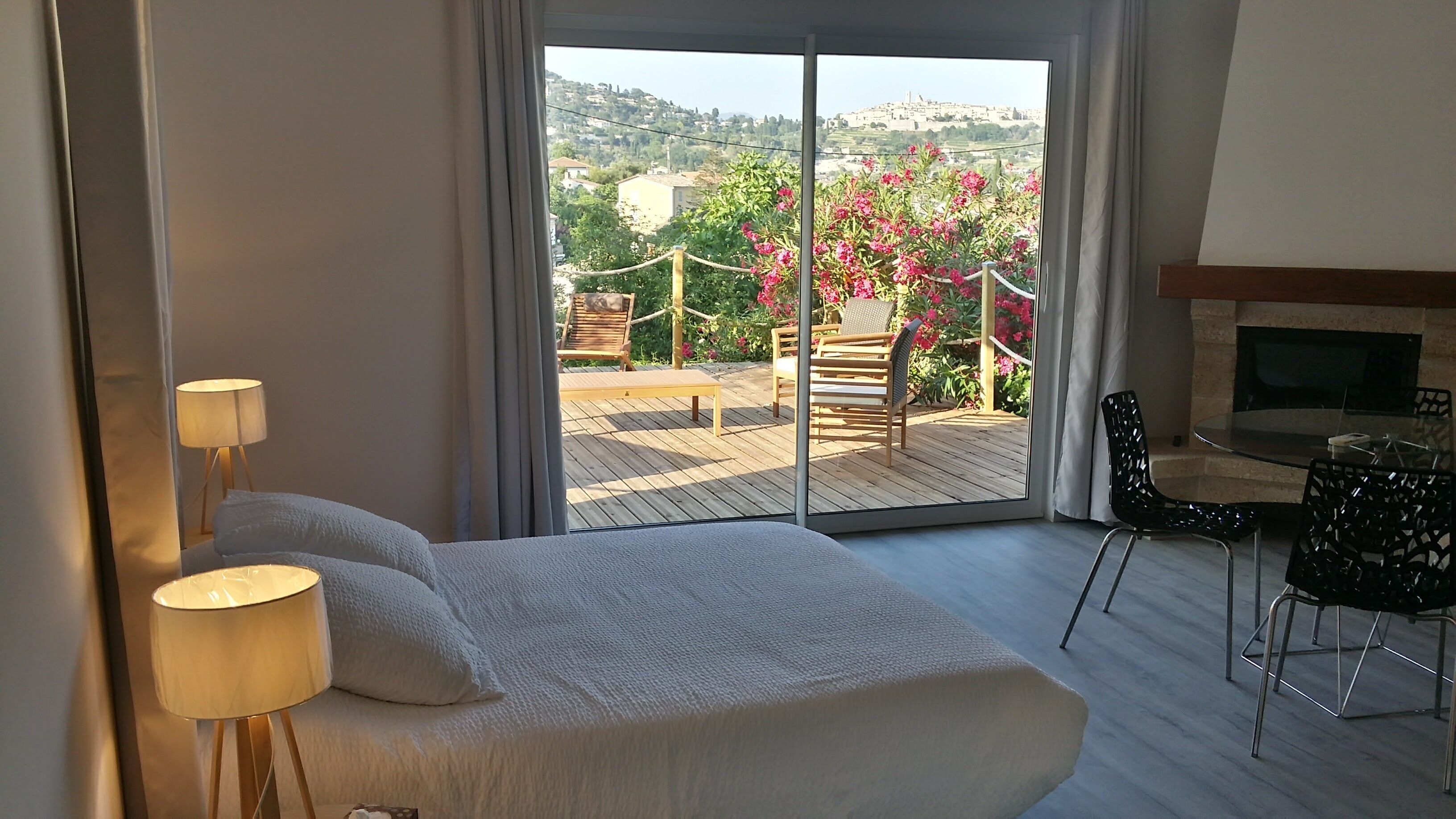 Habitación de hotel con cama, escritorio y vistas a un balcón con mobiliario exterior y un arbusto de flores.