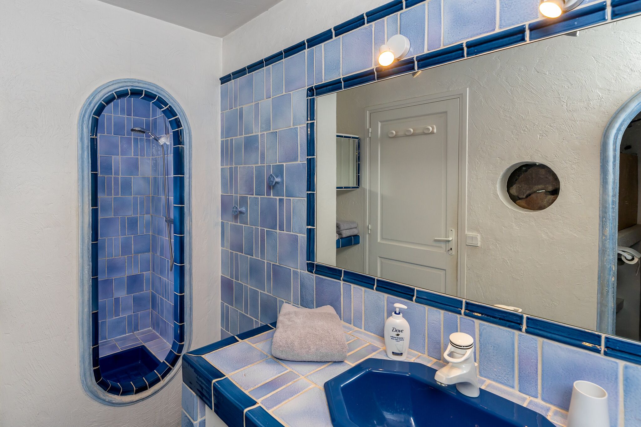A bathroom with a blue-tiled shower, a sink with a towel, and a bottle of Dove soap.