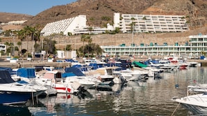 A marina with multiple boats docked, a hotel with a modern design, and a mountainous backdrop.