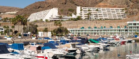A marina with multiple boats docked, a hotel with a modern design, and a mountainous backdrop.