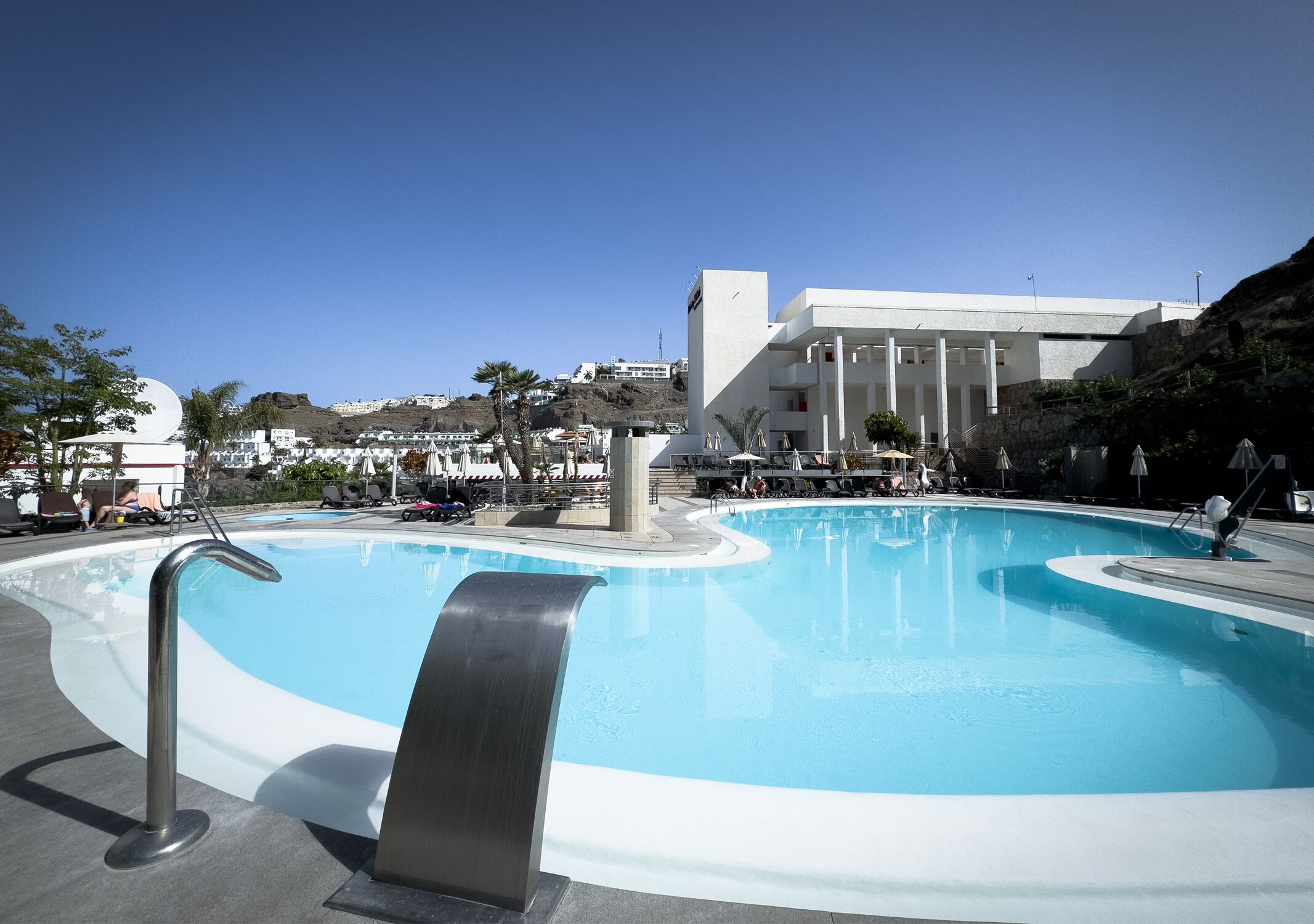 Una piscina de disseny modern, envoltada de gandules i ombrel·les, amb un edifici al fons.