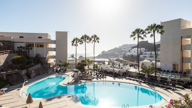 A resort pool area with a large pool, sun loungers, umbrellas, and palm trees.