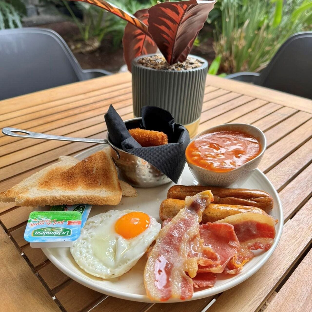 A breakfast plate with toast, fried egg, bacon, sausages, and a small bowl of tomato sauce.