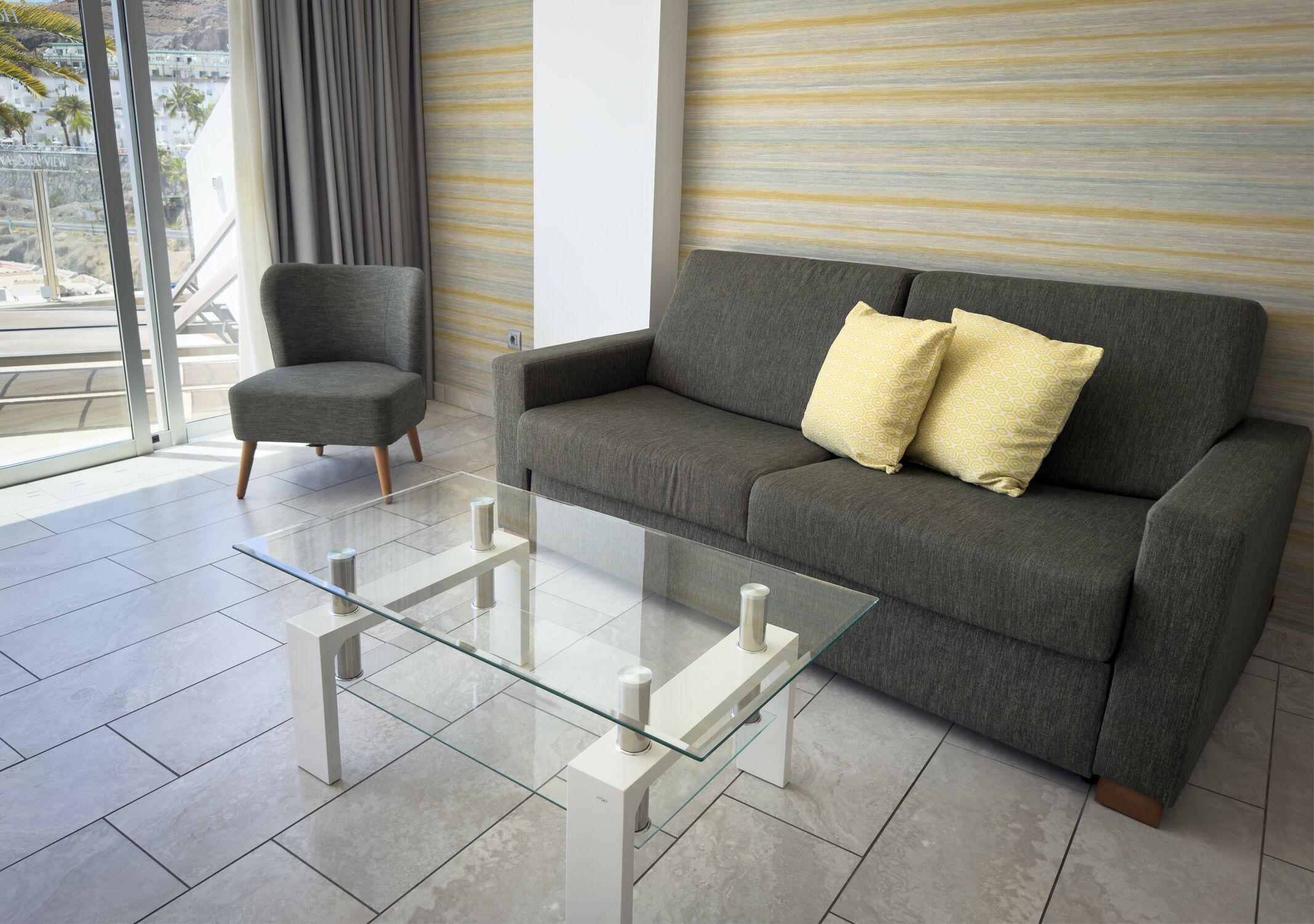 A modern living room with a grey sofa, a glass coffee table, and a grey armchair.