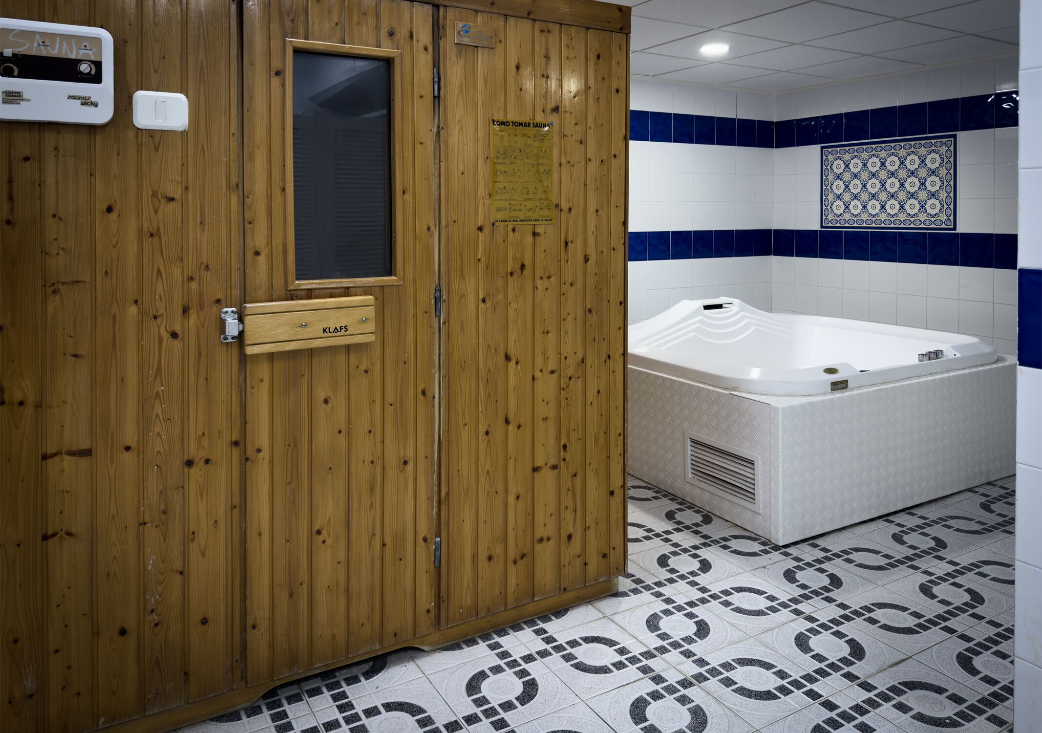 Une porte en bois avec une fenêtre, un panneau indiquant la présence d’un sauna, et une baignoire blanche dans une pièce dont le sol est recouvert de carreaux à motifs.