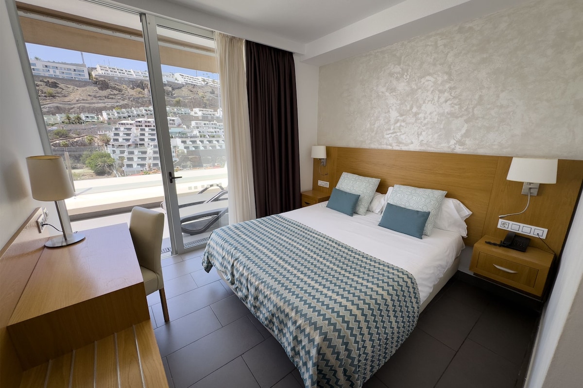 A hotel room with a bed, bedside tables, a desk, and a view of buildings through a window.