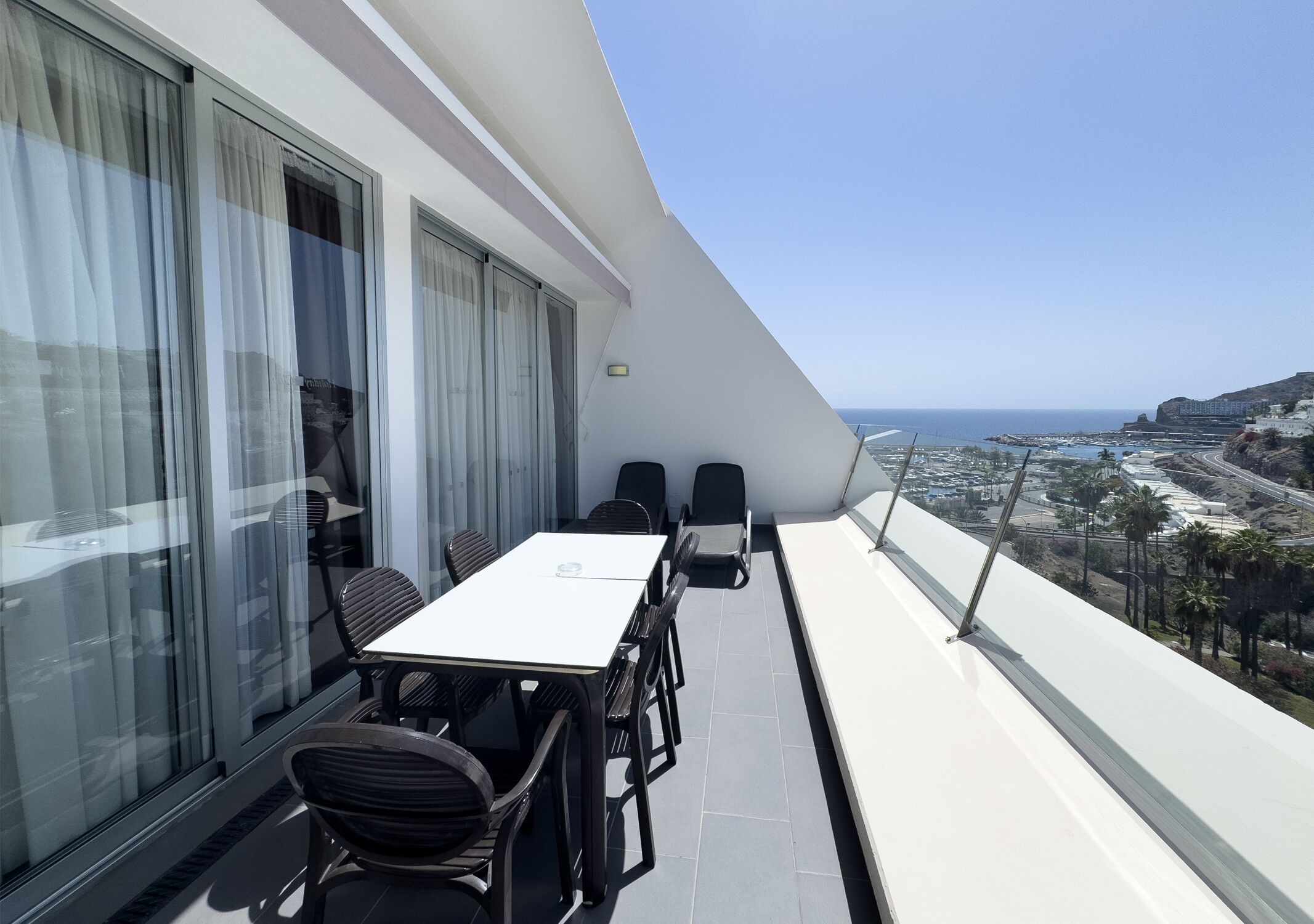 A balcony with a table and chairs, offering a view of the sea and a coastal town.