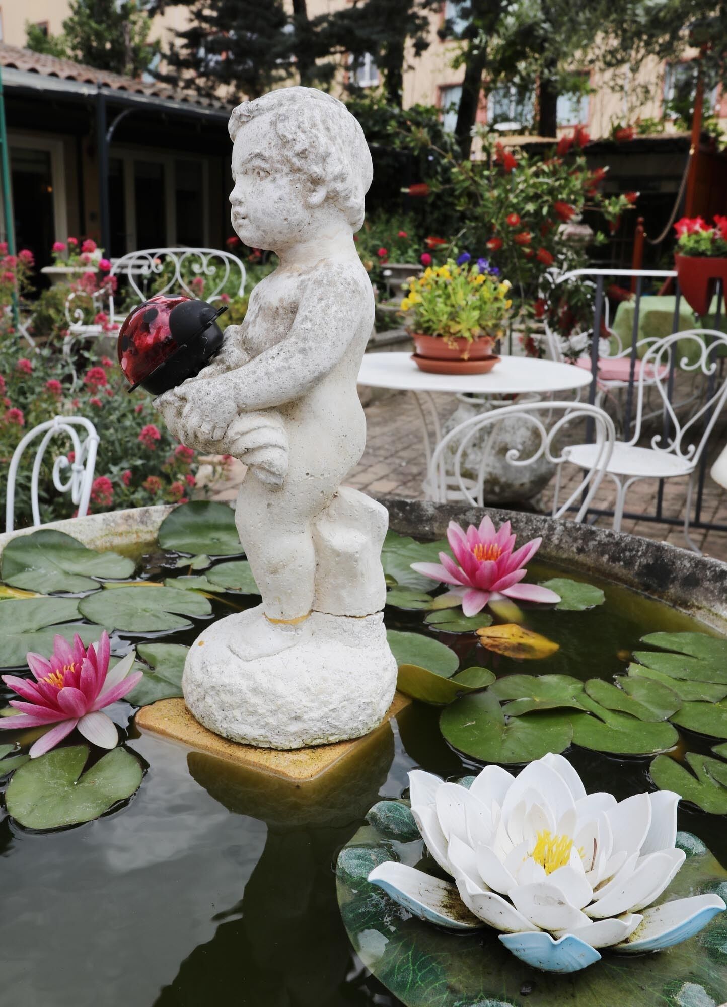 Una estatua de jardín de un niño sosteniendo un objeto rojo, rodeada de lirios acuáticos y plantas en macetas.