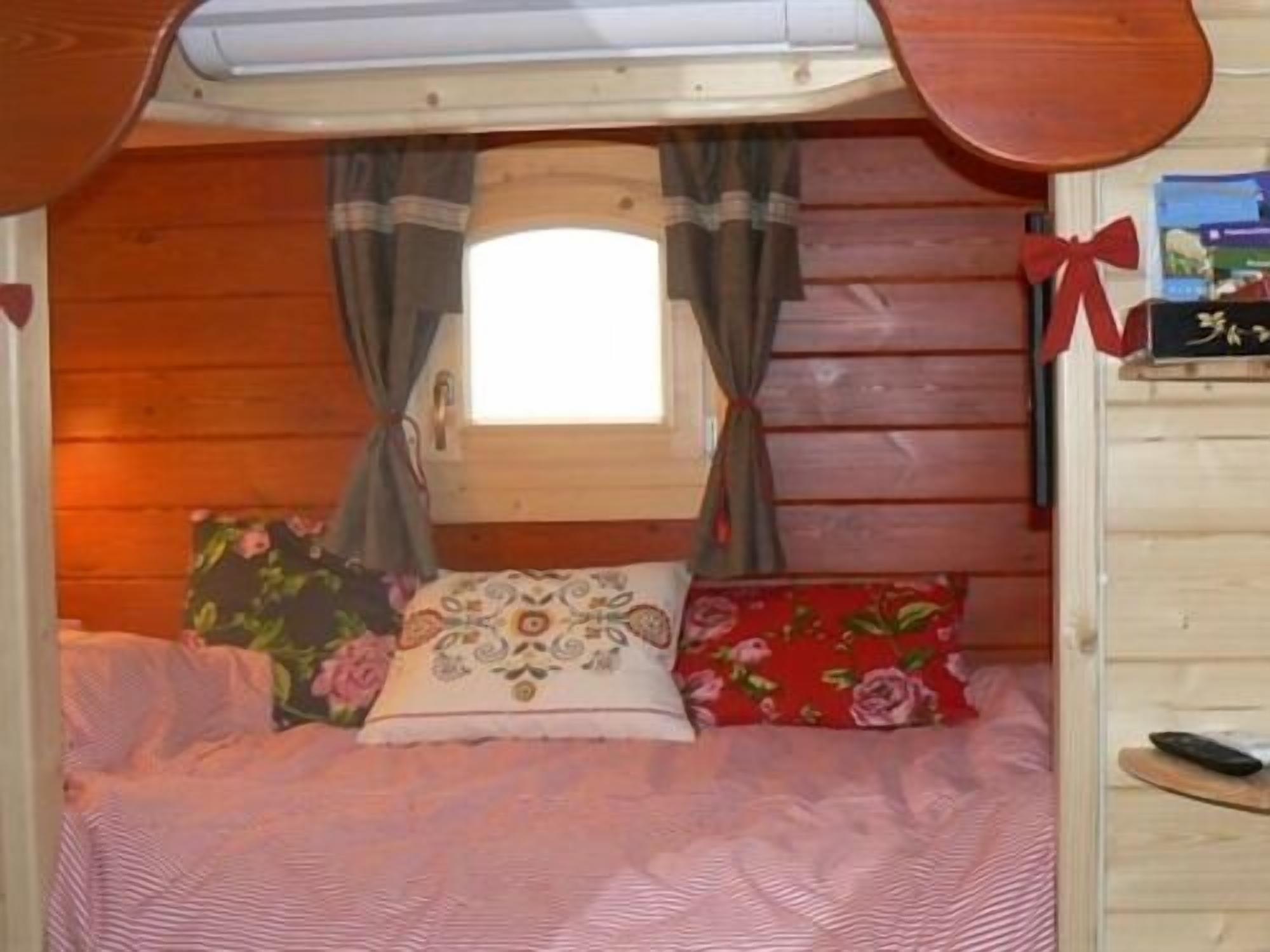 A cozy cabin-style bedroom with a single bed, floral and patterned pillows, a small window with curtains, and a wooden headboard.