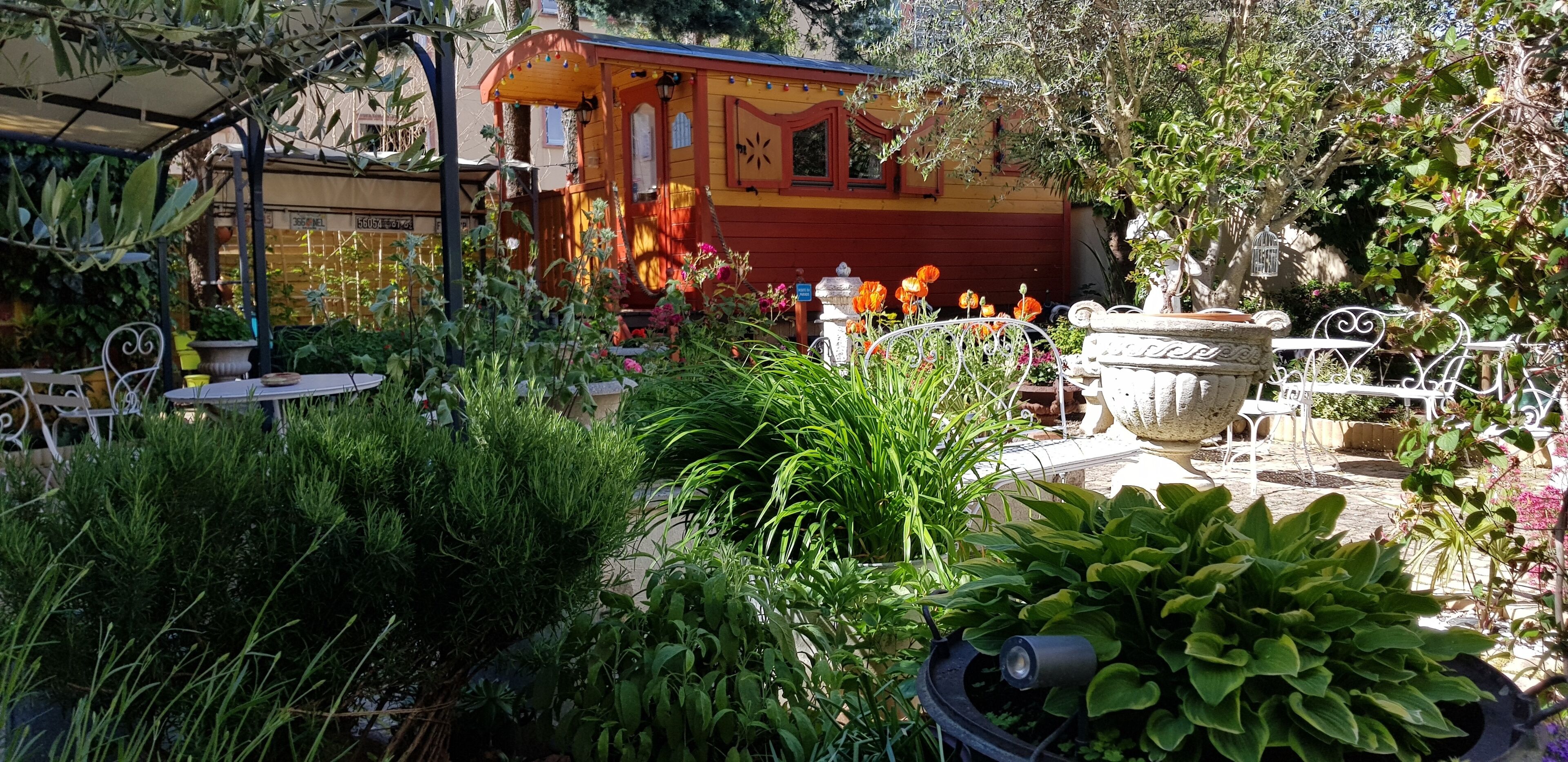 A garden with a small yellow and red building, surrounded by various plants and a metal fence.
