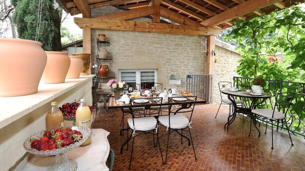 Une terrasse couverte avec un mur en pierre, des poutres en bois, une table dressée pour le repas et des plantes en pot.